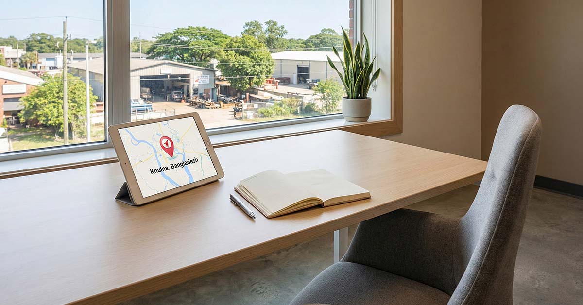 A modern office desk setup with a tablet showing a map pin and a window view of Khalishpur town outside