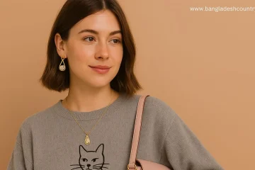 Smiling woman in a grey sweater with a black cat design, featuring gold teardrop earrings and a matching necklace. A pink bag is visible.