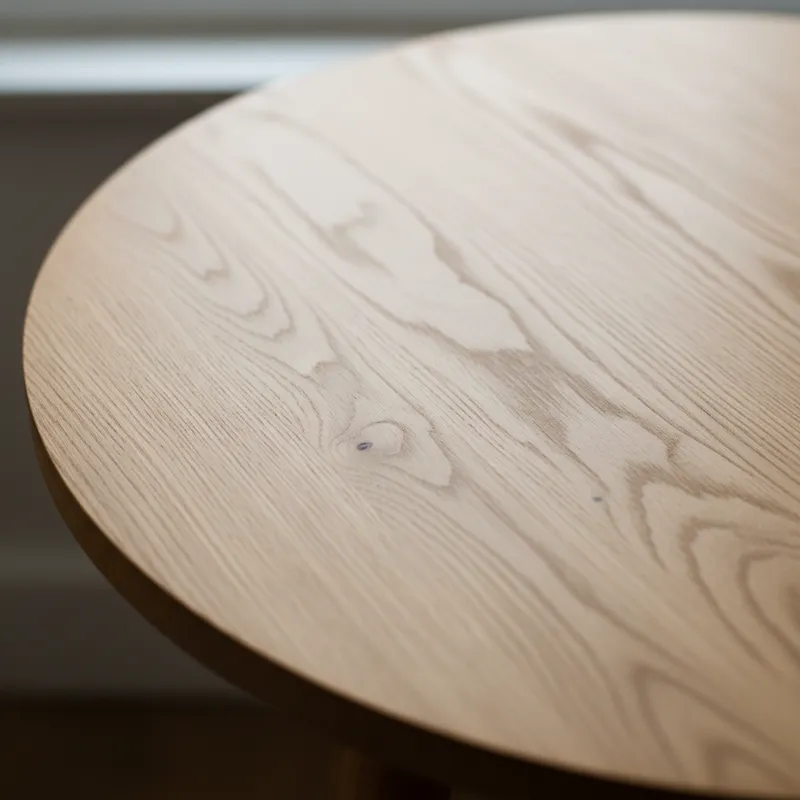 A close-up view of a small circular table's tabletop, showing grain of a light oak wood, with subtle natural imperfections.