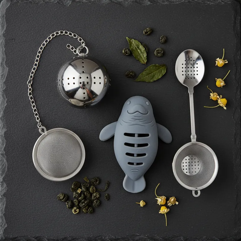 An overhead shot displaying a variety of tea infusers on a dark slate background: a classic mesh ball, a polished perforated steel ball, a fun silicon